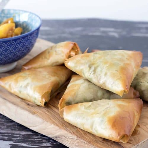 Baked samosas made with filo pastry on wooden board with blue bowl in background