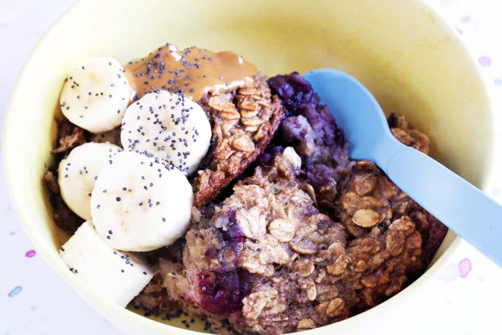 blueberry baked oats in yellow bowl with bananas, nut butter and seeds